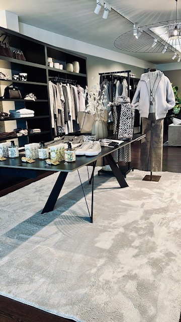 Modern boutique interior with a black display table featuring shoes, candles, and accessories; clothing racks along the wall and a gray hoodie on a mannequin, in a neutral, inviting space.
